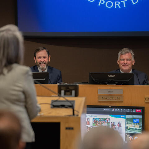 Port CEO and Commissioner smile at a woman at a podium during a Port board meeting