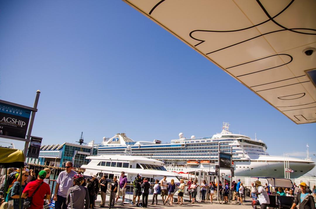 a crowd walks along the North Embarcadero at the Port of San Diego there is a cruise ship in the background.