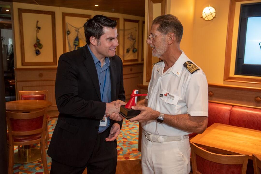 a man in a suit shakes the hand of the captain on a cruise ship. That man is Adam Deaton.