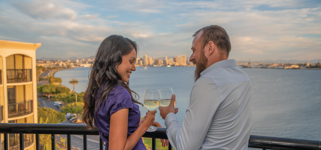 A couple enjoys wine on a hotel balcony overlooking the bay and sunset