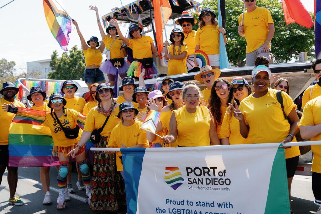 Port employees in yellow shirts and colorful accessories pose behind a Pride Parade banner.