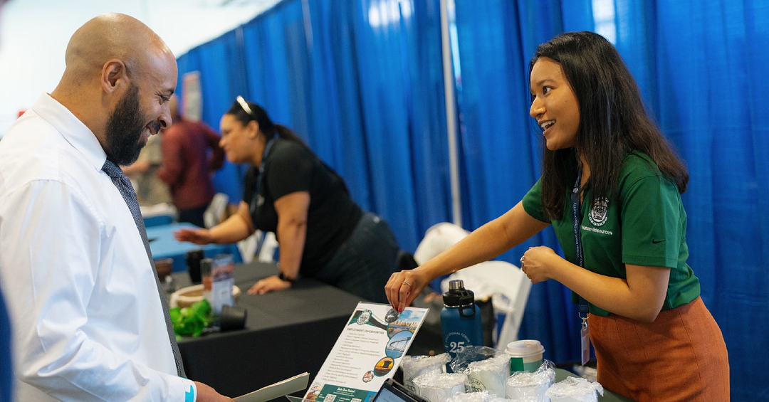 A smiling woman hands a flyer to a bearded man