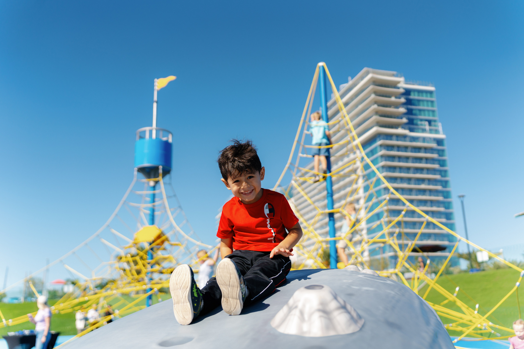Child in red shirt, sitting at Harbor Park Playground in Chula Vista