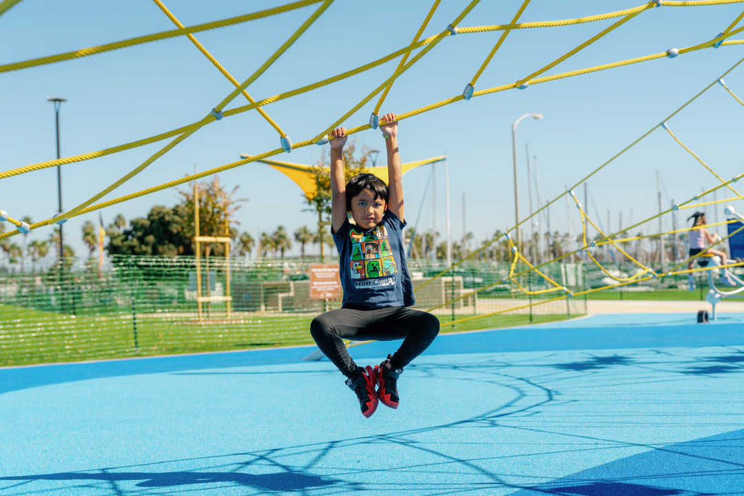 Child in grey shirt, hanging from a play structure in Harbor Park Playground in Chula Vista
