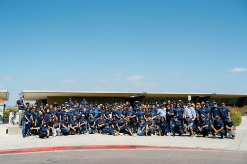 Large group of Port employees in blue t-shirts pose for photo in front of the Living Coast Discovery Center with a blue sky above them.