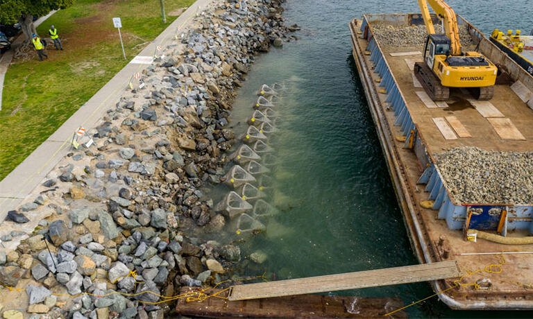 a barge with a crane sits to the right of the shore, triangular concrete blocks are also along the shore