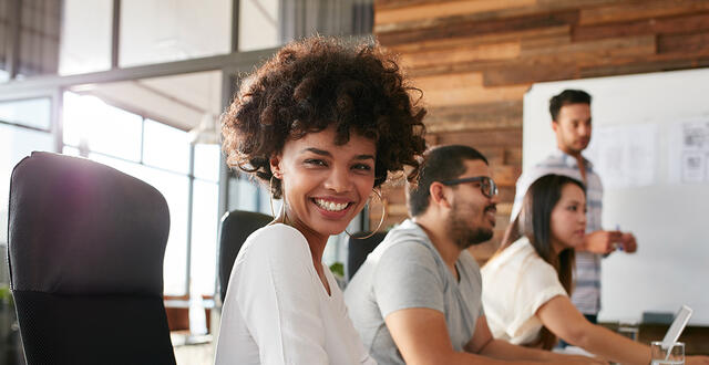 a Black woman smiles at the camera - colleagues in the background