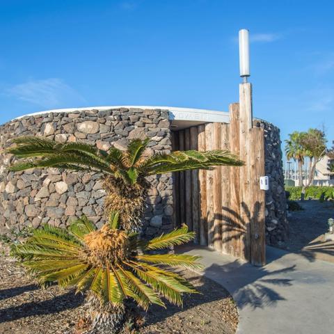 The restroom at Harbor Island Park. A round, rock covered structure with a path and palm trees.
