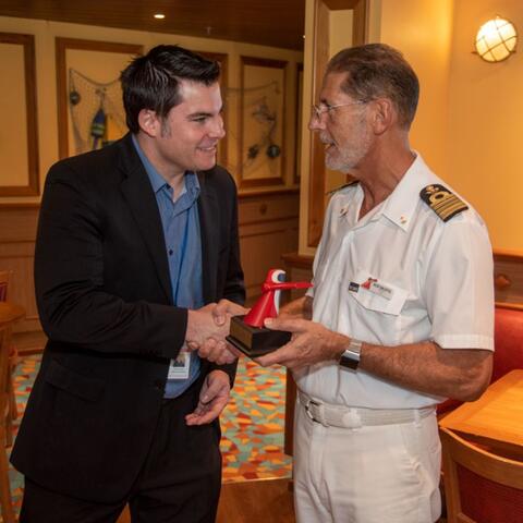 a man in a suit shakes the hand of the captain on a cruise ship. That man is Adam Deaton.