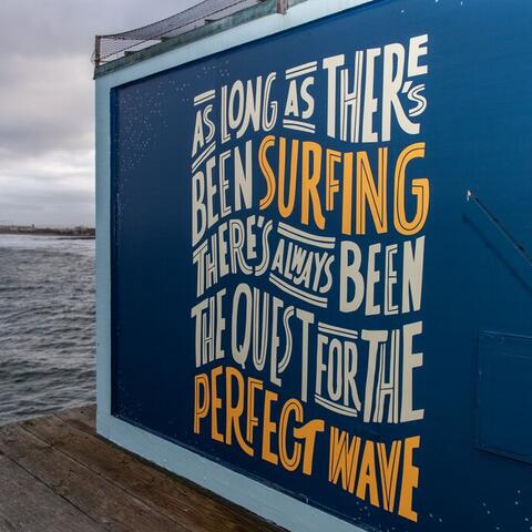 Colorful mural on the Imperial Beach Pier that says, "As long as there's been surfing there's always been the quest for the perfect wave."