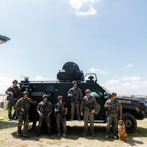 7 people in green uniforms and helmets pose with a dog in front of a vehicle